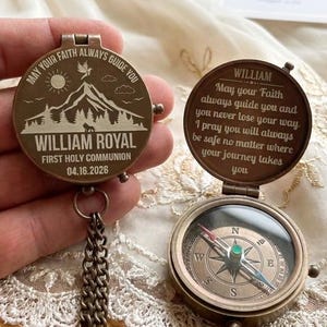 May include: A bronze-colored compass with a hinged lid, featuring engraved text and a detailed mountain scene. The compass includes a chain and a glass-covered dial with directional markings. The text reads "May your Faith always guide you" and "William Royal, First Holy Communion 04.16.2026."