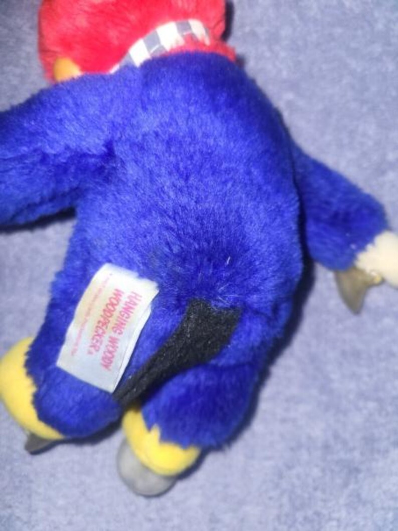 May include: A plush Woody Woodpecker toy, primarily blue with a red head and yellow feet. The toy has a black tail and a white tag that reads "Hanging Woody Woodpecker."