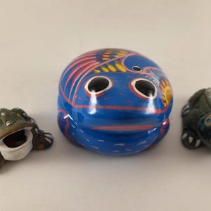 May include: Three ceramic frog figurines. A large blue frog with red and yellow accents, a small green frog with many eyes, and a small green frog with white spots.