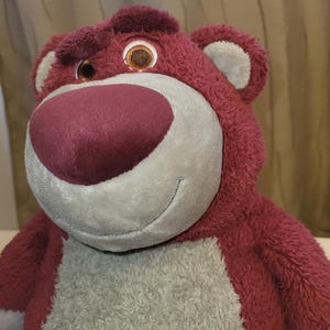 May include: A soft toy bear with red-pink fur, a large pink nose, and a smiling face. The bear has brown eyes and grey accents on the muzzle and chest. The toy is designed to be soft and cuddly, suitable for children.