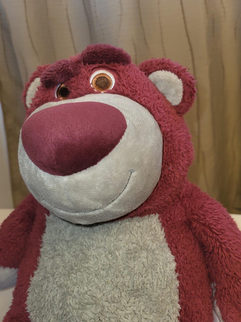 May include: A soft toy bear with red-pink fur, a large pink nose, and a smiling face. The bear has brown eyes and grey accents on the muzzle and chest. The toy is designed to be soft and cuddly, suitable for children.