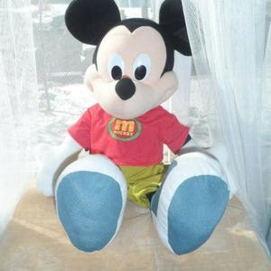May include: A plush Mickey Mouse doll with large black ears, a red shirt with a green and yellow emblem, yellow trousers, and large blue and white feet. The doll is sitting on a light brown surface.