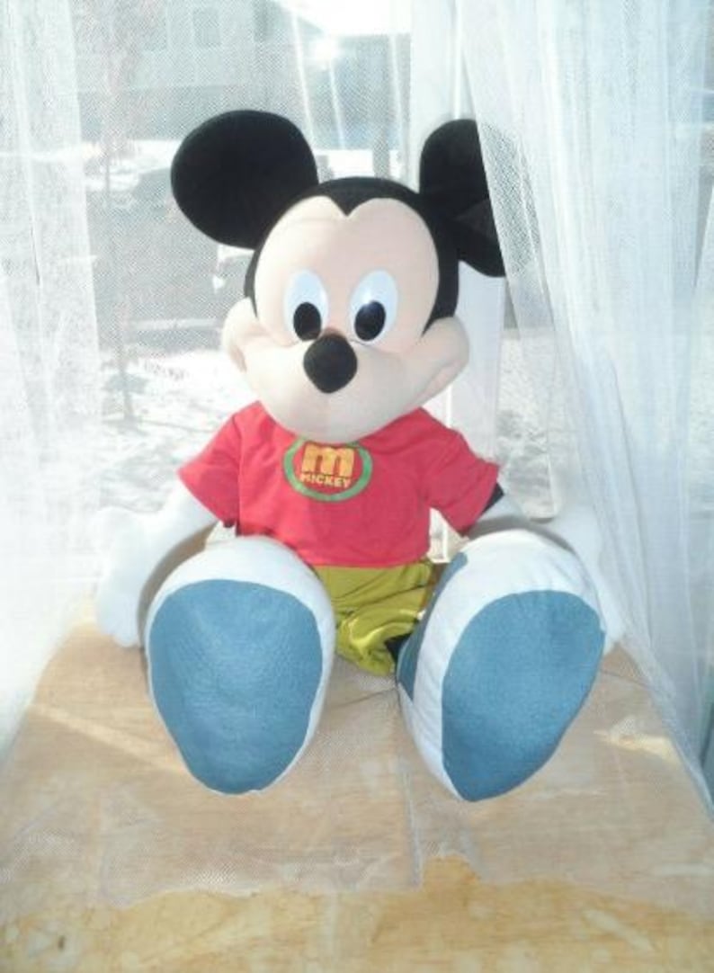 May include: A plush Mickey Mouse doll with large black ears, a red shirt with a green and yellow emblem, yellow trousers, and large blue and white feet. The doll is sitting on a light brown surface.