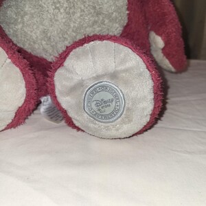 May include: A soft plush toy with a light grey body and dark pink details. The foot of the toy has a circular patch with the text "Genuine Original Disney Store Authentic". The toy is made of a fuzzy material.