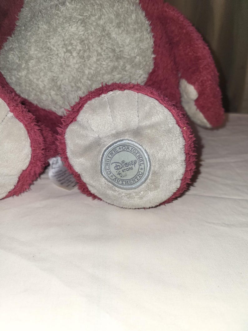 May include: A soft plush toy with a light grey body and dark pink details. The foot of the toy has a circular patch with the text "Genuine Original Disney Store Authentic". The toy is made of a fuzzy material.