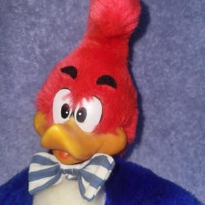 May include: A plush Woody Woodpecker doll with a red head, yellow beak, and a blue and white striped bow tie. The doll has white eyes and black eyebrows, with blue and white striped bow tie.