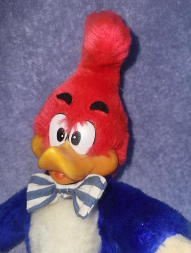 May include: A plush Woody Woodpecker doll with a red head, yellow beak, and a blue and white striped bow tie. The doll has white eyes and black eyebrows, with blue and white striped bow tie.