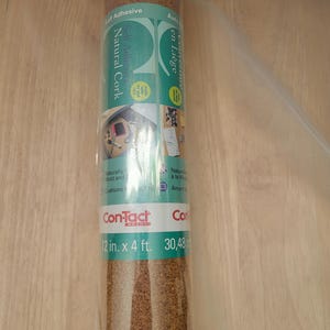 May include: A roll of self-adhesive natural cork, 12 inches by 4 feet. The label includes the brand name "Con-Tact" and text in English and French. The cork has a brown, textured appearance and is rolled up.