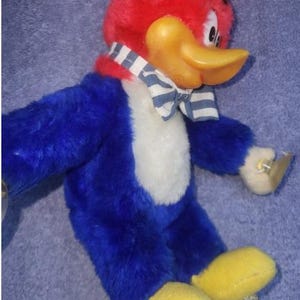 May include: A plush Woody Woodpecker toy. The toy has a red head and tuft, a yellow beak, and blue body with a white chest. It wears a striped bow tie and has yellow feet. The toy is a classic cartoon character.