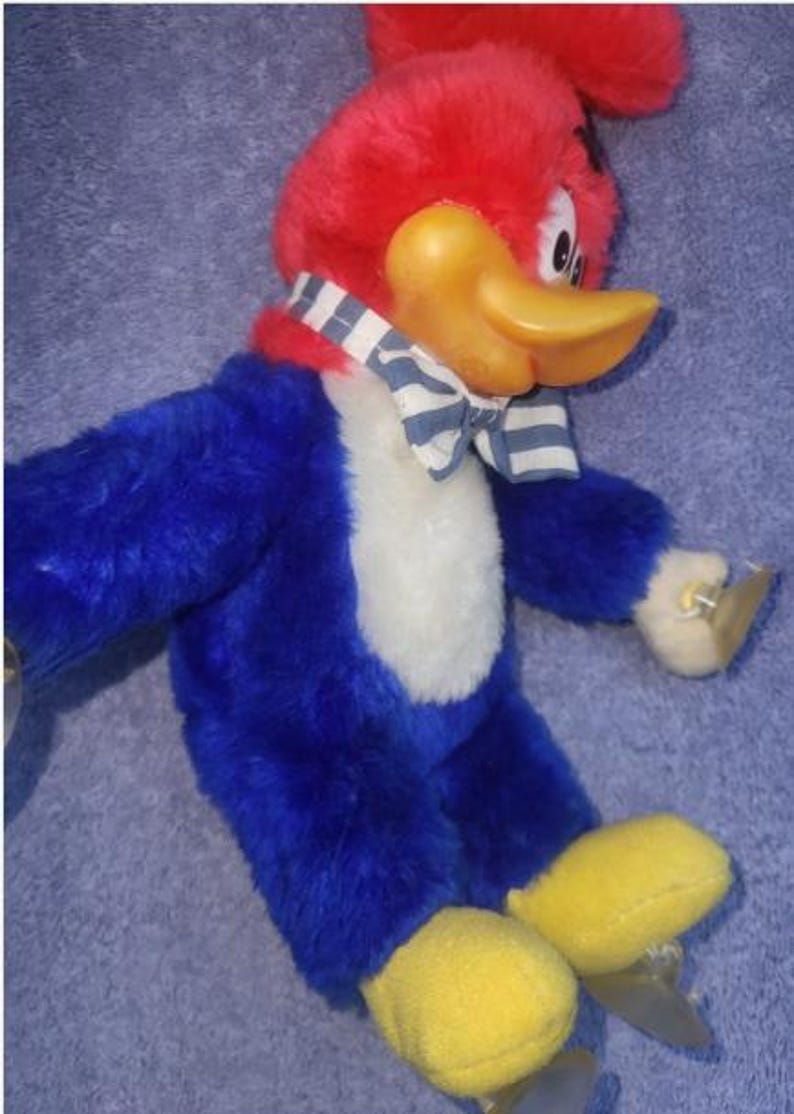 May include: A plush Woody Woodpecker toy. The toy has a red head and tuft, a yellow beak, and blue body with a white chest. It wears a striped bow tie and has yellow feet. The toy is a classic cartoon character.