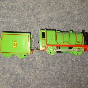 May include: A green toy steam locomotive with a matching tender featuring the number 3 in yellow. The train has red detailing, black wheels, and a silver coupling hook. The engine body displays red lining and a black front section.