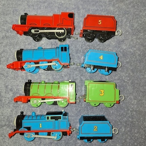 May include: A collection of four toy train engines and their matching tenders. The set includes a red engine with tender number 5, a blue engine with tender number 4, a green engine with tender number 3, and a blue engine with tender number 1 and tender number 2.