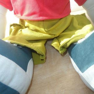 May include: A close-up of a doll wearing a red shirt with the word "MICKEY" in green lettering, yellow shorts, and blue and white striped trousers. The doll is sitting on a beige surface.