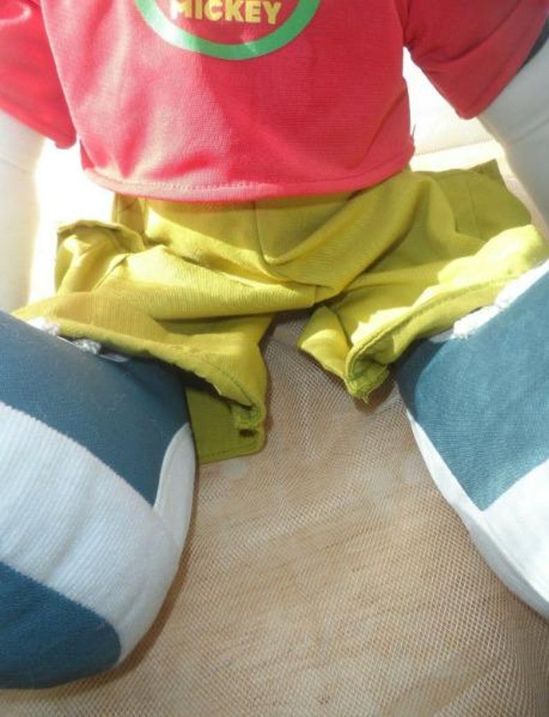 May include: A close-up of a doll wearing a red shirt with the word "MICKEY" in green lettering, yellow shorts, and blue and white striped trousers. The doll is sitting on a beige surface.