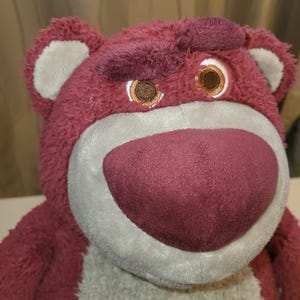 May include: A maroon plush bear toy with a light grey muzzle and a maroon nose. The bear has brown eyes and a furrowed brow, giving it a slightly grumpy expression. The toy is soft and cuddly.