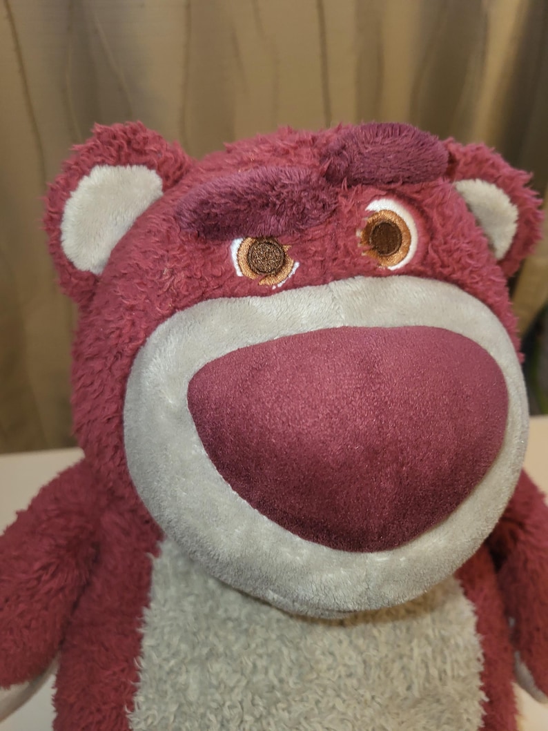 May include: A maroon plush bear toy with a light grey muzzle and a maroon nose. The bear has brown eyes and a furrowed brow, giving it a slightly grumpy expression. The toy is soft and cuddly.