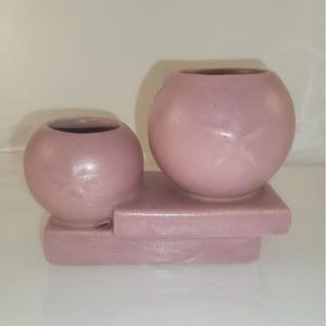 May include: Two pink ceramic planters with a rectangular base. The larger planter is round and the smaller planter is slightly oval shaped.