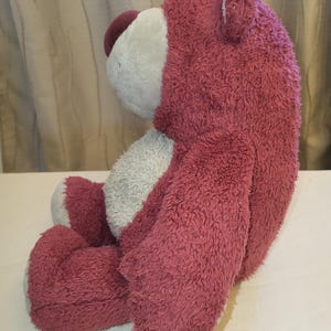 May include: A pink, seated teddy bear with a fuzzy texture. The bear has a white face and belly, with a darker purple nose and head. The toy is designed to be seated, with its legs and arms positioned.