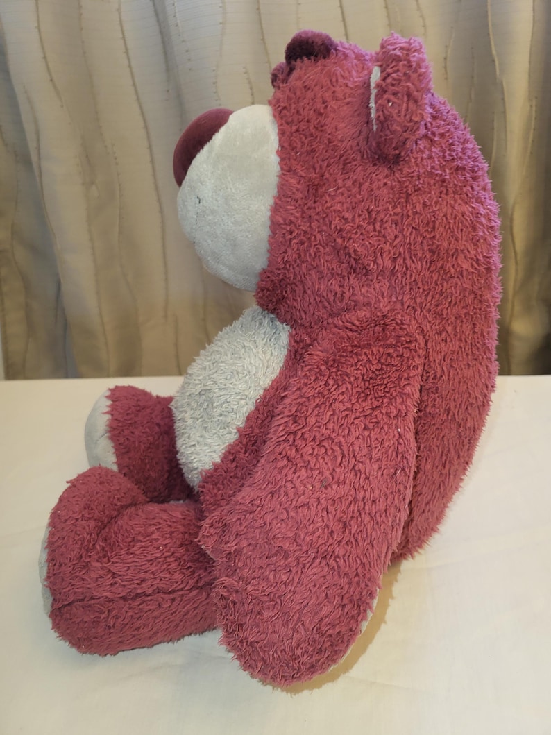 May include: A pink, seated teddy bear with a fuzzy texture. The bear has a white face and belly, with a darker purple nose and head. The toy is designed to be seated, with its legs and arms positioned.