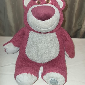 May include: A berry-coloured plush bear with a grey belly, snout, and foot pads. The bear has a friendly expression and small, brown eyes. A Disney logo is visible on one foot.
