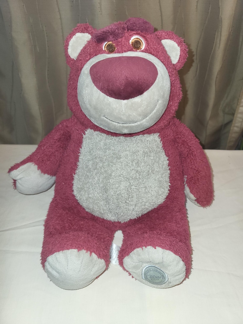 May include: A berry-coloured plush bear with a grey belly, snout, and foot pads. The bear has a friendly expression and small, brown eyes. A Disney logo is visible on one foot.