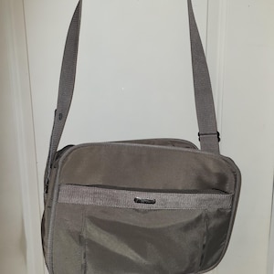 May include: A gray messenger bag with a shoulder strap and a handle. The bag has a rectangular shape with a front pocket and a zippered compartment. The bag is hanging on a door.
