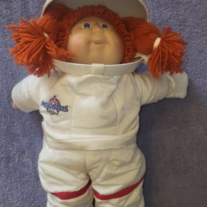 May include: A vintage Cabbage Patch Kids astronaut doll. The doll has a white astronaut suit with red accents and the text "Astronauts". The doll's head is inside a white helmet. The doll has orange yarn hair.