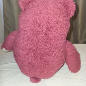 May include: A pink teddy bear toy with a soft, textured surface. The bear has rounded ears, a small tail, and white accents on its paws. The toy is placed on a white surface.