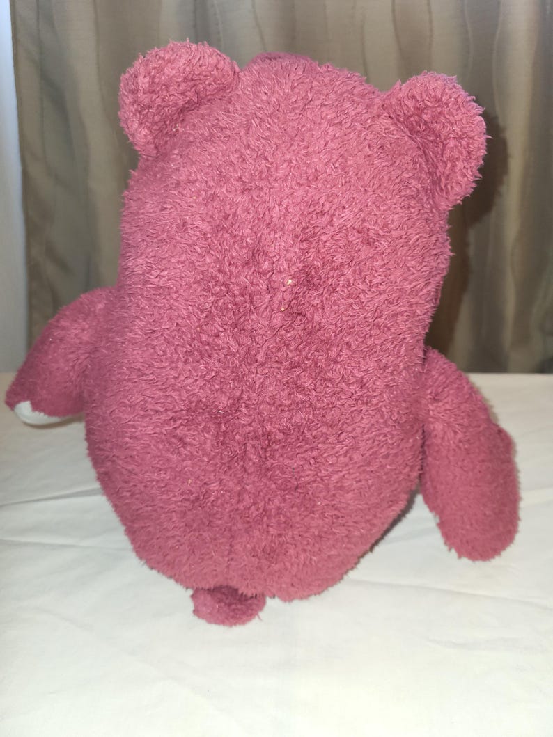 May include: A pink teddy bear toy with a soft, textured surface. The bear has rounded ears, a small tail, and white accents on its paws. The toy is placed on a white surface.