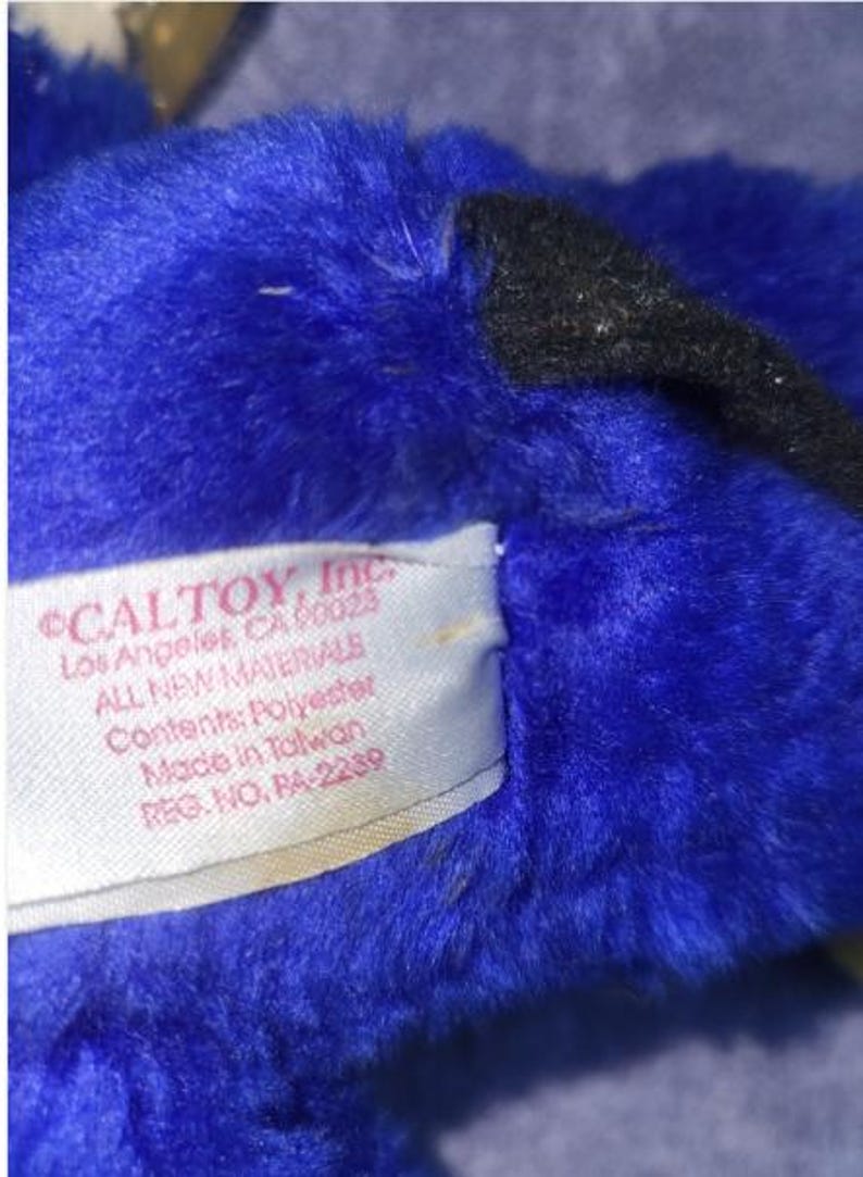 May include: Close-up of a plush toy with vibrant blue fur and a black accent. A tag reads "CALTOY, Inc. Los Angeles, CA 90023 ALL NEW MATERIALS Contents: Polyester Made in Taiwan REG. NO. RA-2239".