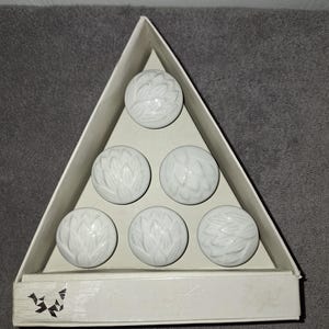 May include: A set of six white ceramic knobs with a raised, embossed floral petal pattern, arranged in a triangular formation inside a matching triangular cardboard box. The box features a small black graphic logo on the bottom left corner.