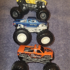 May include: Three toy monster trucks are displayed. The top truck is yellow with a bull design and the word "Bulldozer". The middle truck is blue with "Bigfoot" on the side. The bottom truck is orange with "Bad Company" on the side.