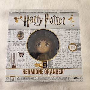 May include: A boxed Hermione Granger vinyl figure from the Harry Potter series. The box features the Harry Potter logo, a Hogwarts crest, and the text "Hermione Granger". The figure is visible through a circular window.