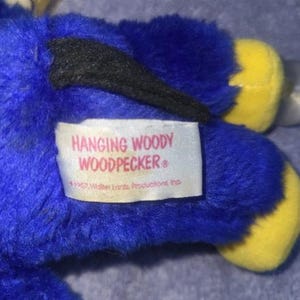 May include: A plush toy of Woody Woodpecker, primarily blue with yellow feet and a black wing. The toy has a white beak and a tag that reads "Hanging Woody Woodpecker".