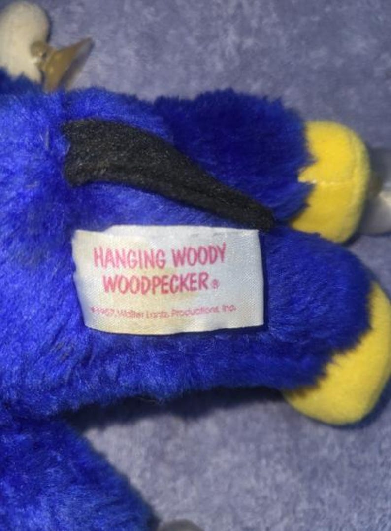 May include: A plush toy of Woody Woodpecker, primarily blue with yellow feet and a black wing. The toy has a white beak and a tag that reads "Hanging Woody Woodpecker".