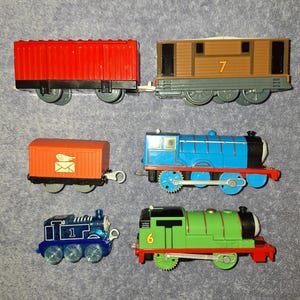May include: A collection of six toy trains and train cars. The set includes a red freight car, a brown engine numbered 7, an orange mail car with a letter icon, a blue engine, a small translucent blue engine numbered 1, and a green engine numbered 6.