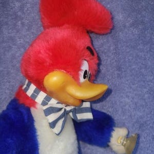 May include: A plush Woody Woodpecker doll. The bird has a red head and crest, a yellow beak, and a blue and white striped bow tie. The doll is wearing a blue and white striped scarf and a blue jacket.