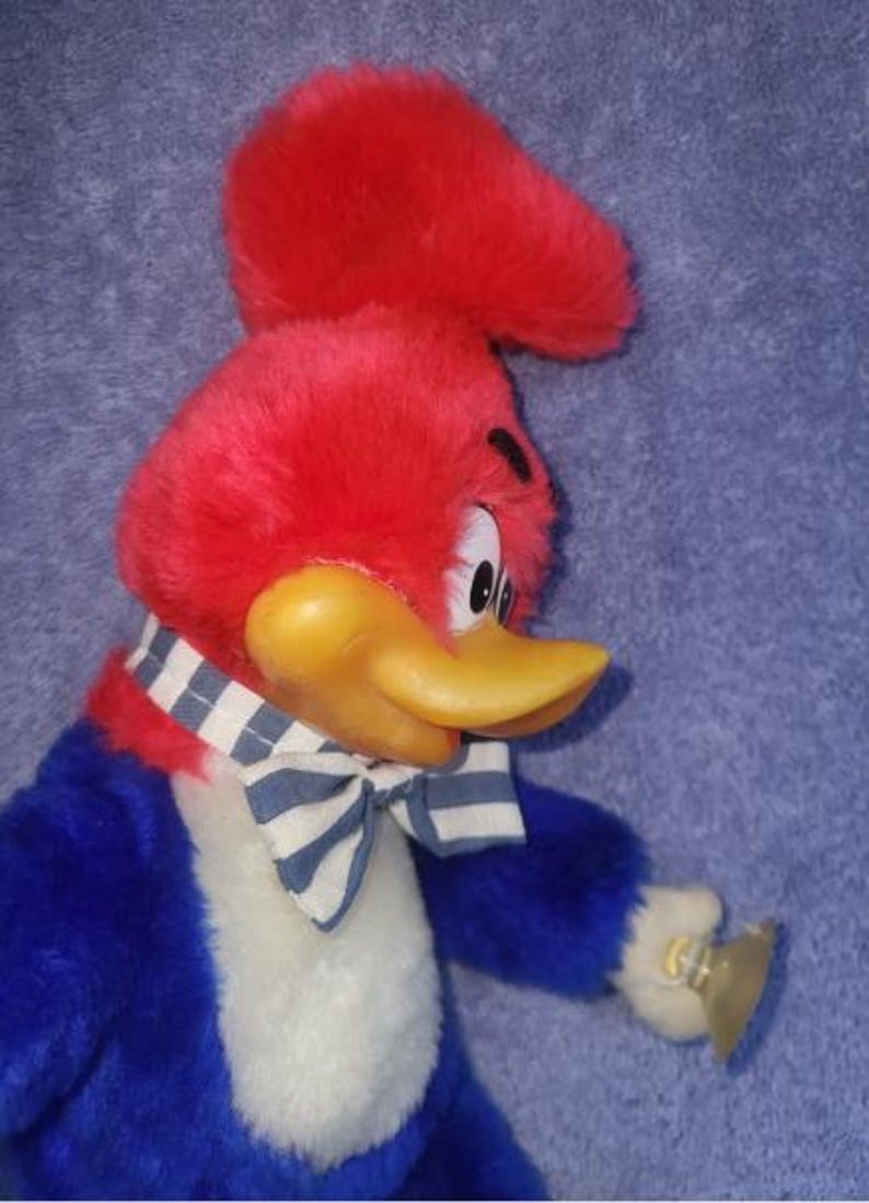 May include: A plush Woody Woodpecker doll. The bird has a red head and crest, a yellow beak, and a blue and white striped bow tie. The doll is wearing a blue and white striped scarf and a blue jacket.
