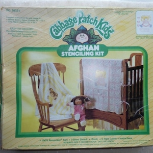 May include: A Cabbage Patch Kids Afghan Stenciling Kit with a wooden rocking chair and a crib. The kit includes yarn, a stencil, a brush, six paint colors, and instructions. The kit requires a size G crochet hook, which is not included.