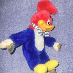 May include: A plush Woody Woodpecker toy. The toy has a red head with a tuft, a yellow beak, and a blue body with white chest. It wears a blue and white striped bow tie and has yellow feet with metal skates.