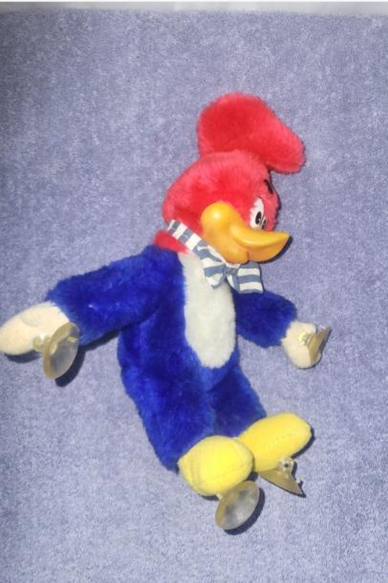 May include: A plush Woody Woodpecker toy. The toy has a red head with a tuft, a yellow beak, and a blue body with white chest. It wears a blue and white striped bow tie and has yellow feet with metal skates.