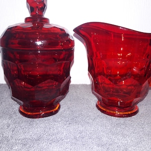 May include: A set of two pieces of red glass tableware consisting of a lidded sugar bowl and a creamer pitcher. Both items feature a faceted, geometric pattern across their surfaces, a circular base, and a deep, translucent red color throughout.