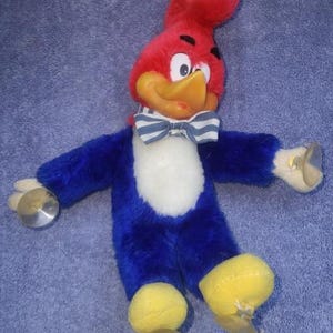 May include: A plush toy of Woody Woodpecker, featuring a red head, yellow beak, and blue body. The toy has a white chest, a striped bow tie, and yellow feet. It holds cymbals in each hand, and is a vintage collectible.