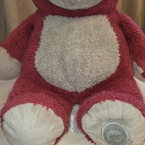 May include: A red bear-shaped plush toy with a light grey belly, face, and feet. The toy has a smiling expression and a Disney Store authentic tag on one foot.