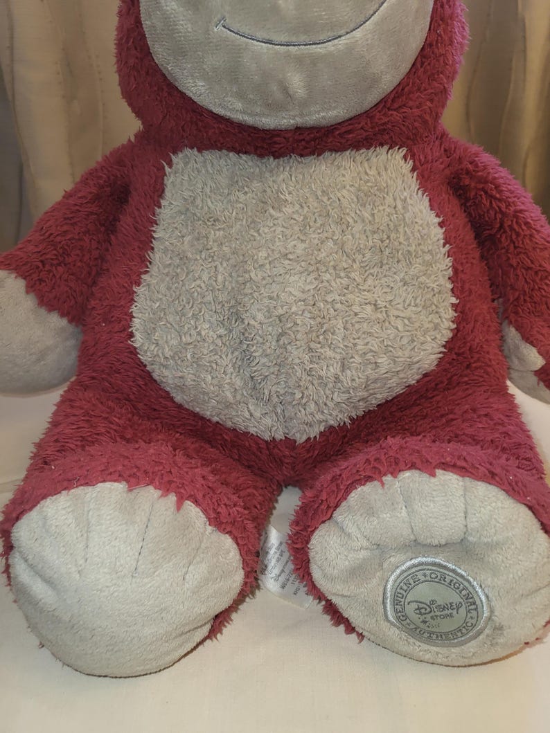 May include: A red bear-shaped plush toy with a light grey belly, face, and feet. The toy has a smiling expression and a Disney Store authentic tag on one foot.
