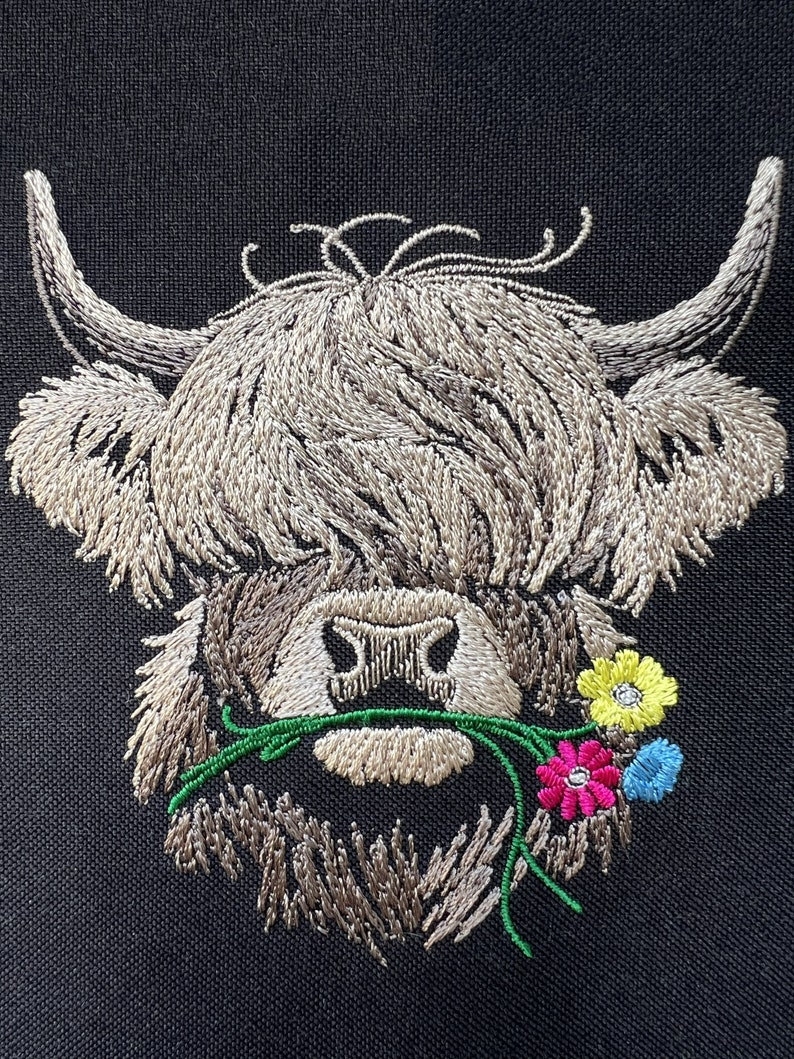 May include: Embroidered design of a Highland cow with brown fur, a white face, and a green stem with pink, yellow, and blue flowers in its mouth.