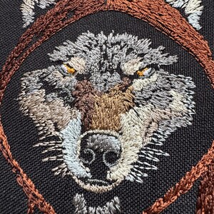 K&ouml;nnte beinhalten: Nahaufnahme eines gestickten Wolfskopfes mit braunem, grauem und wei&szlig;em Garn. Die Augen des Wolfes sind leuchtend orange. Das Gesicht des Wolfes ist von einem braunen gestickten Kreis umgeben.