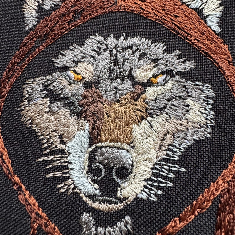 K&ouml;nnte beinhalten: Nahaufnahme eines gestickten Wolfskopfes mit braunem, grauem und wei&szlig;em Garn. Die Augen des Wolfes sind leuchtend orange. Das Gesicht des Wolfes ist von einem braunen gestickten Kreis umgeben.
