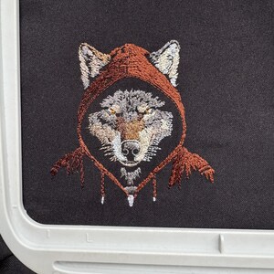K&ouml;nnte beinhalten: Gestickte Darstellung eines Wolfes, der ein braunes Kapuzenpullover tr&auml;gt. Das Fell des Wolfes ist grau und wei&szlig;, und seine Augen sind gelb.