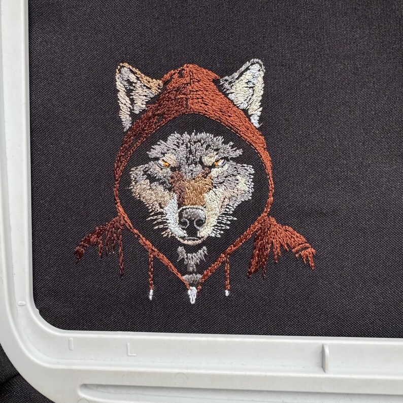 K&ouml;nnte beinhalten: Gestickte Darstellung eines Wolfes, der ein braunes Kapuzenpullover tr&auml;gt. Das Fell des Wolfes ist grau und wei&szlig;, und seine Augen sind gelb.